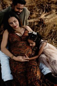 A pregnant woman in a patterned dress sits outdoors, smiling with a man behind her. A young girl in a light dress hugs and kisses the woman’s bump affectionately—capturing a tender moment for their Adelaide baby photos among the dry grass.