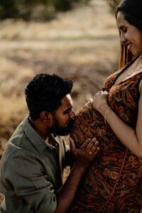 A man kneels and kisses a woman’s pregnant bump as she smiles, both looking happy and serene outdoors in natural light—capturing the joy of Adelaide baby photos.