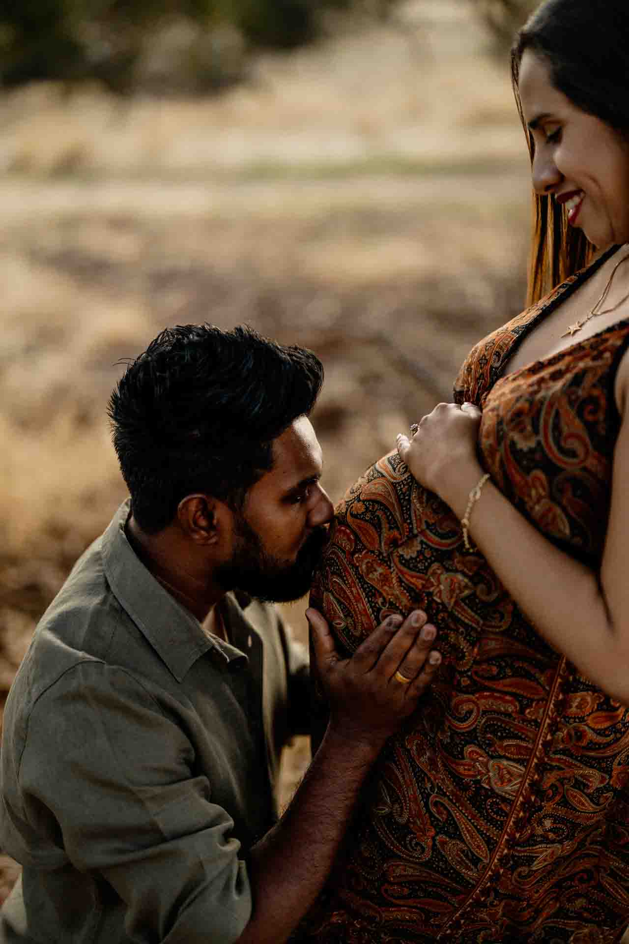 A man kneels and kisses a woman’s pregnant bump as she smiles, both looking happy and serene outdoors in natural light—capturing the joy of Adelaide baby photos.