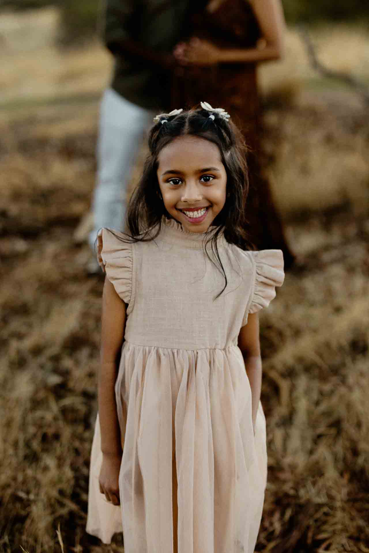 A young girl with long dark hair and butterfly hair clips wears a pale pink dress and smiles at the camera, standing outdoors on dry grass—capturing the sweet charm often seen in Adelaide baby photographs. Two adults are blurred in the background.