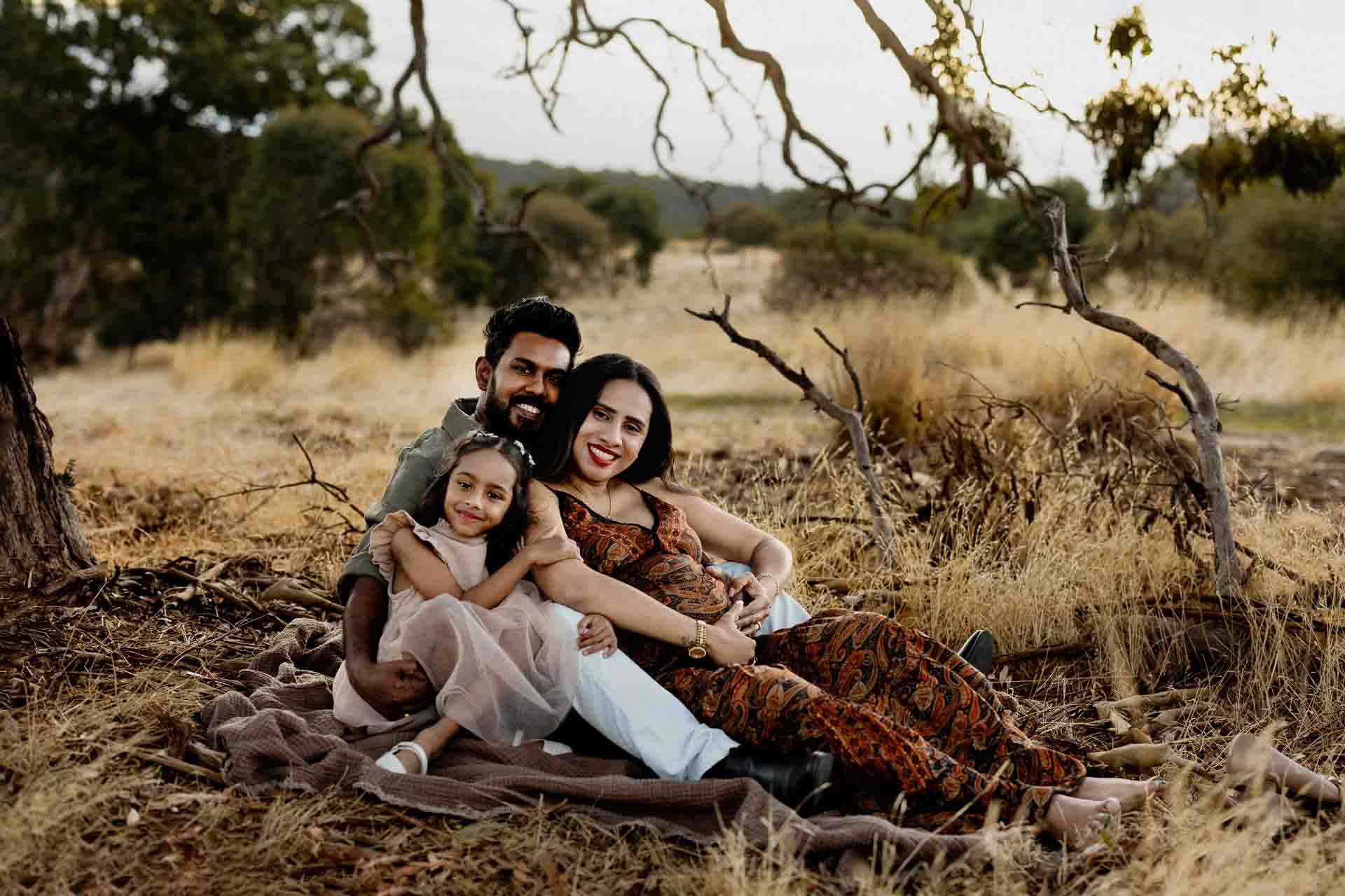 A smiling family of three sits on a blanket outdoors in a dry, grassy area with trees in the background. The parents sit close together as their young daughter hugs them, capturing the warmth typical of Adelaide baby photographs. All appear happy and relaxed.