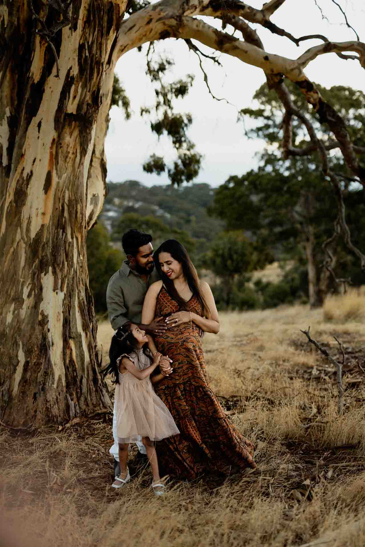 A family of three stands outdoors by a large tree, the father hugging the mother from behind as she smiles, while their young daughter touches her mother’s pregnant tummy—a beautiful moment captured for Adelaide baby photos.