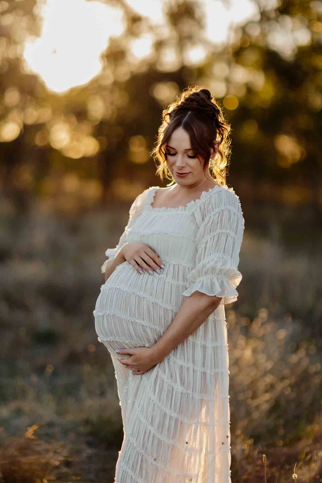 A pregnant mother in a white dress looking at her tummy at a maternity session in Adelaide.