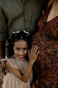 A smiling young girl with a bow in her hair hugs a pregnant woman, gently resting her hand on the woman’s patterned dress and baby bump—capturing a sweet moment in these Adelaide baby photos. An adult stands behind them, partly visible.