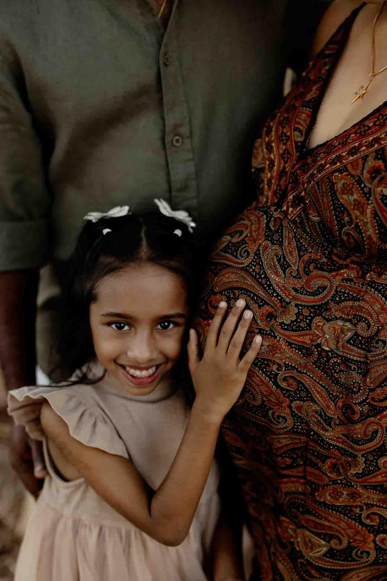 A smiling young girl with a bow in her hair hugs a pregnant woman, gently resting her hand on the woman’s patterned dress and baby bump—capturing a sweet moment in these Adelaide baby photos. An adult stands behind them, partly visible.