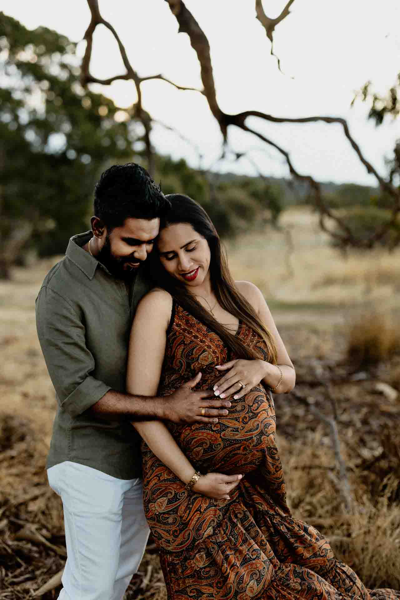 A couple stands outdoors in a grassy field. The man hugs the woman from behind, resting his hands on her pregnant bump. Both appear happy and peaceful, capturing a tender moment perfect for Adelaide baby photos.