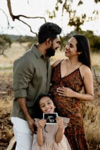 A smiling family stands outdoors; a pregnant woman in a patterned dress embraces her partner whilst their young daughter holds a scan photo, capturing the joy of Adelaide baby photos together.