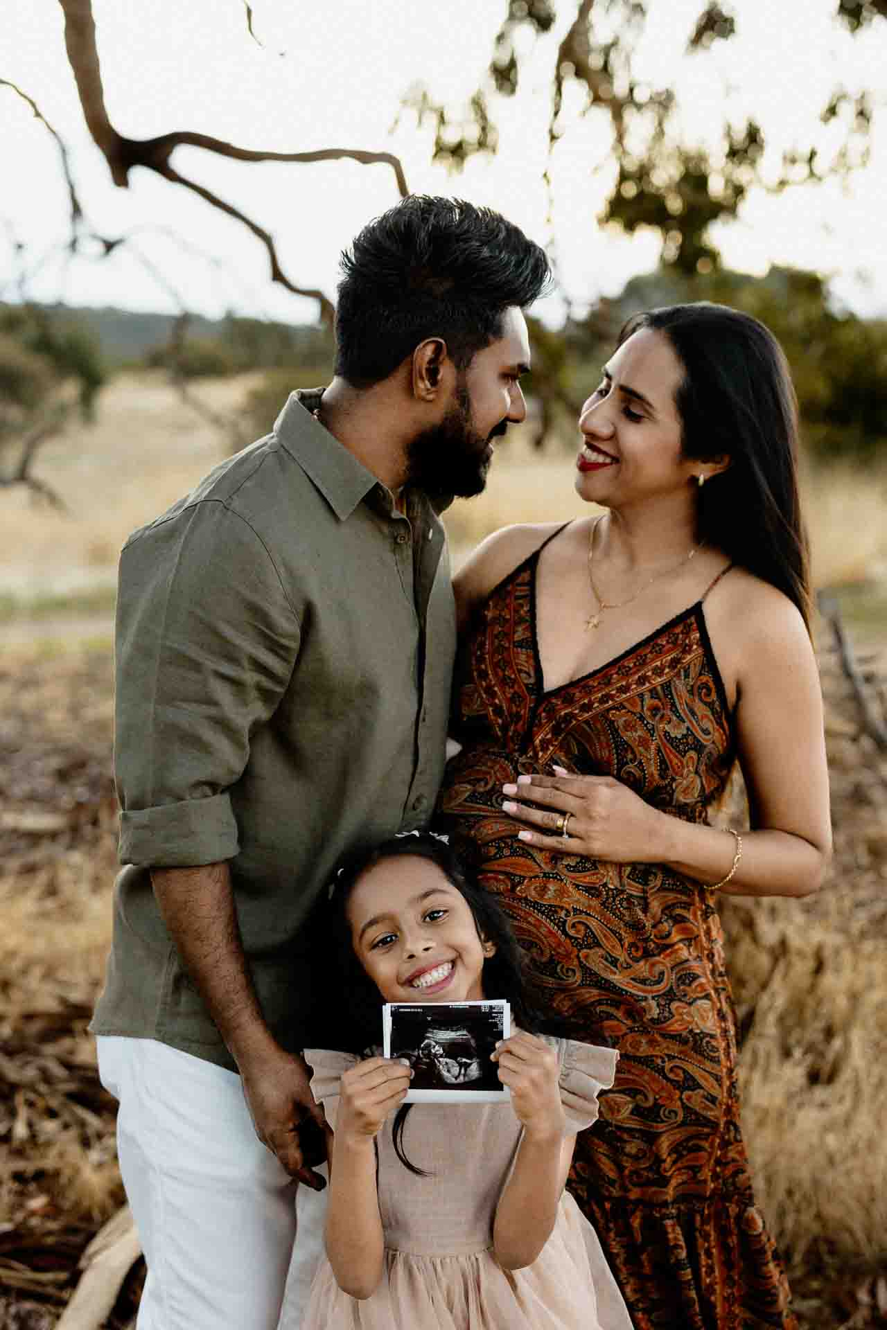 A smiling family stands outdoors; a pregnant woman in a patterned dress embraces her partner whilst their young daughter holds a scan photo, capturing the joy of Adelaide baby photos together.