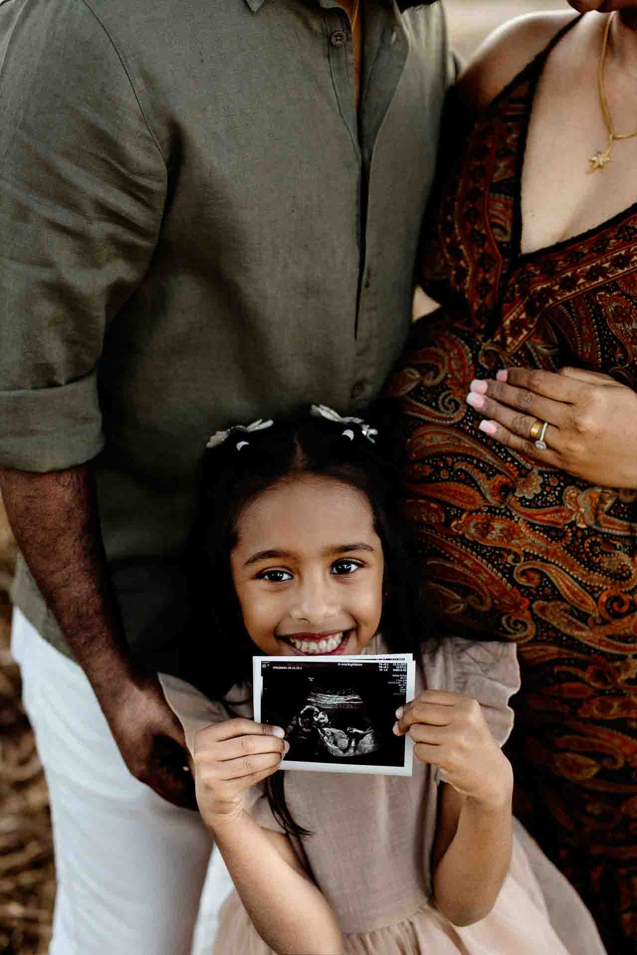 A smiling young girl holds a scan photo while standing in front of a man and a pregnant woman cradling her bump. The happy family shares a warm moment, perfect for Adelaide baby photos.