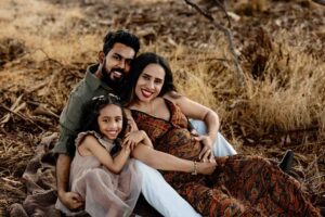 A smiling family of three sits outdoors on dry grass, with the father and pregnant mother sitting close together and their young daughter cuddling in front—all looking happy and relaxed, perfect for Adelaide baby photos.