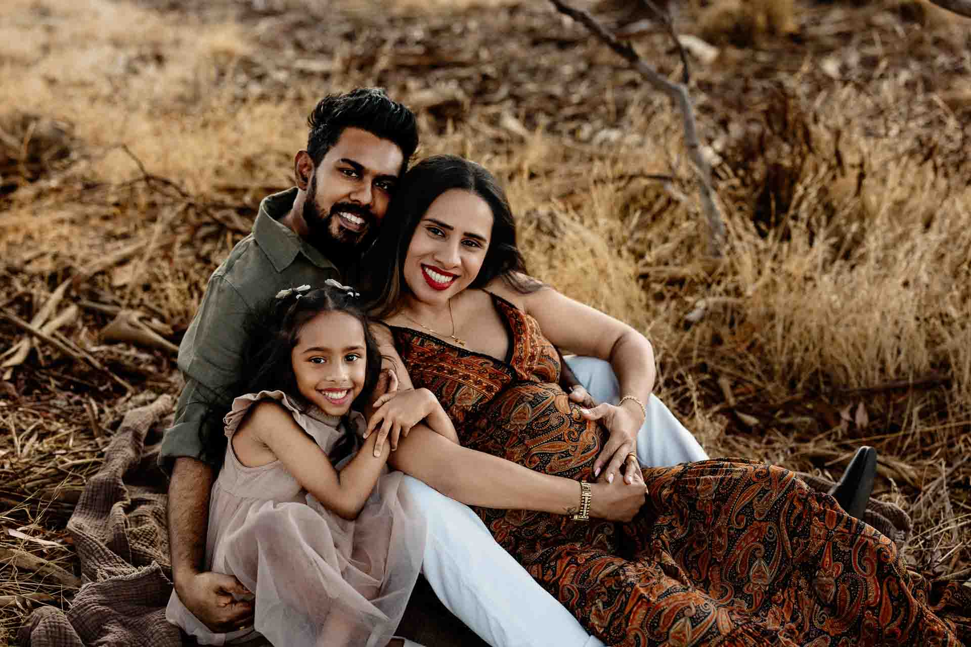 A smiling family of three sits outdoors on dry grass, with the father and pregnant mother sitting close together and their young daughter cuddling in front—all looking happy and relaxed, perfect for Adelaide baby photos.