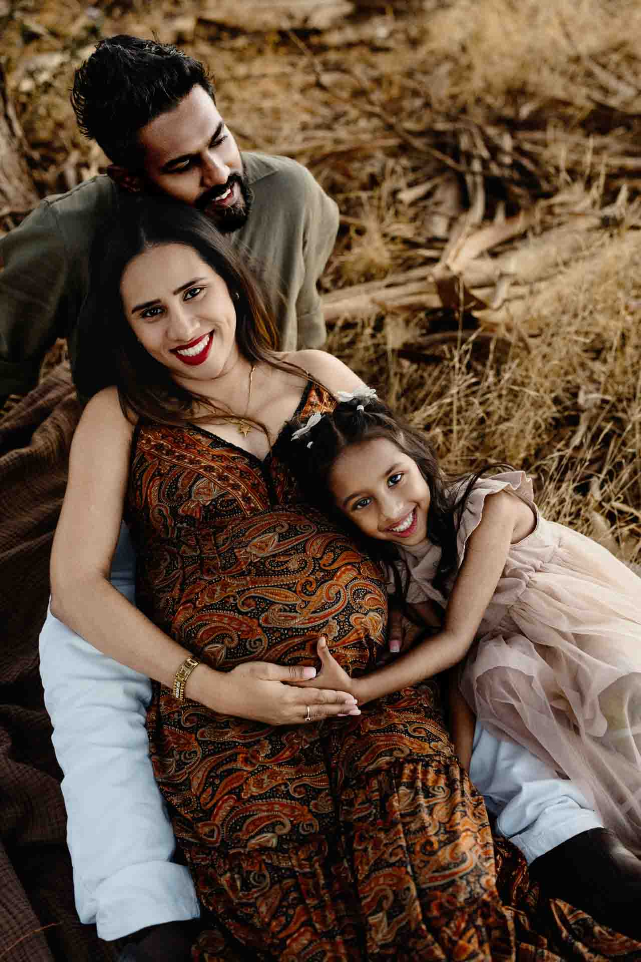 A smiling pregnant woman sits on a blanket outdoors, holding her bump. Capturing precious Adelaide baby photos, a man sits behind her while a young girl in a dress hugs her, all appearing happy and relaxed in a grassy, natural setting.
