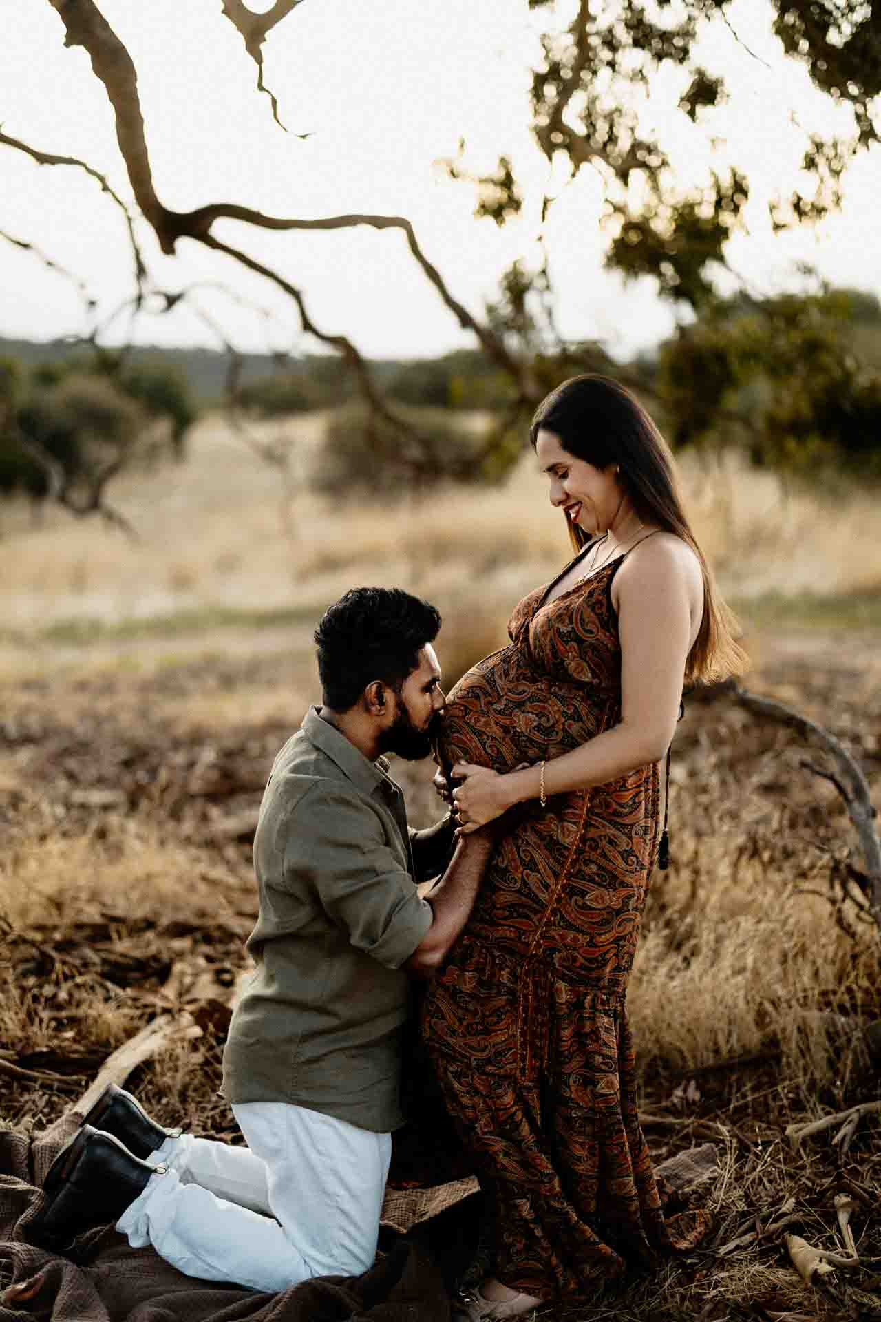 A man kneels outdoors, gently kissing the pregnant belly of a smiling woman standing before him. Surrounded by dry grass and trees in a rustic setting, this tender moment beautifully captures Adelaide baby photos in nature.