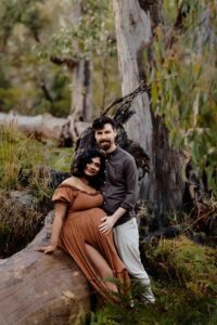 A pregnant mother wearing a rust dress sitting on a fallen tree with her partner at a maternity session in the Adelaide Hills.