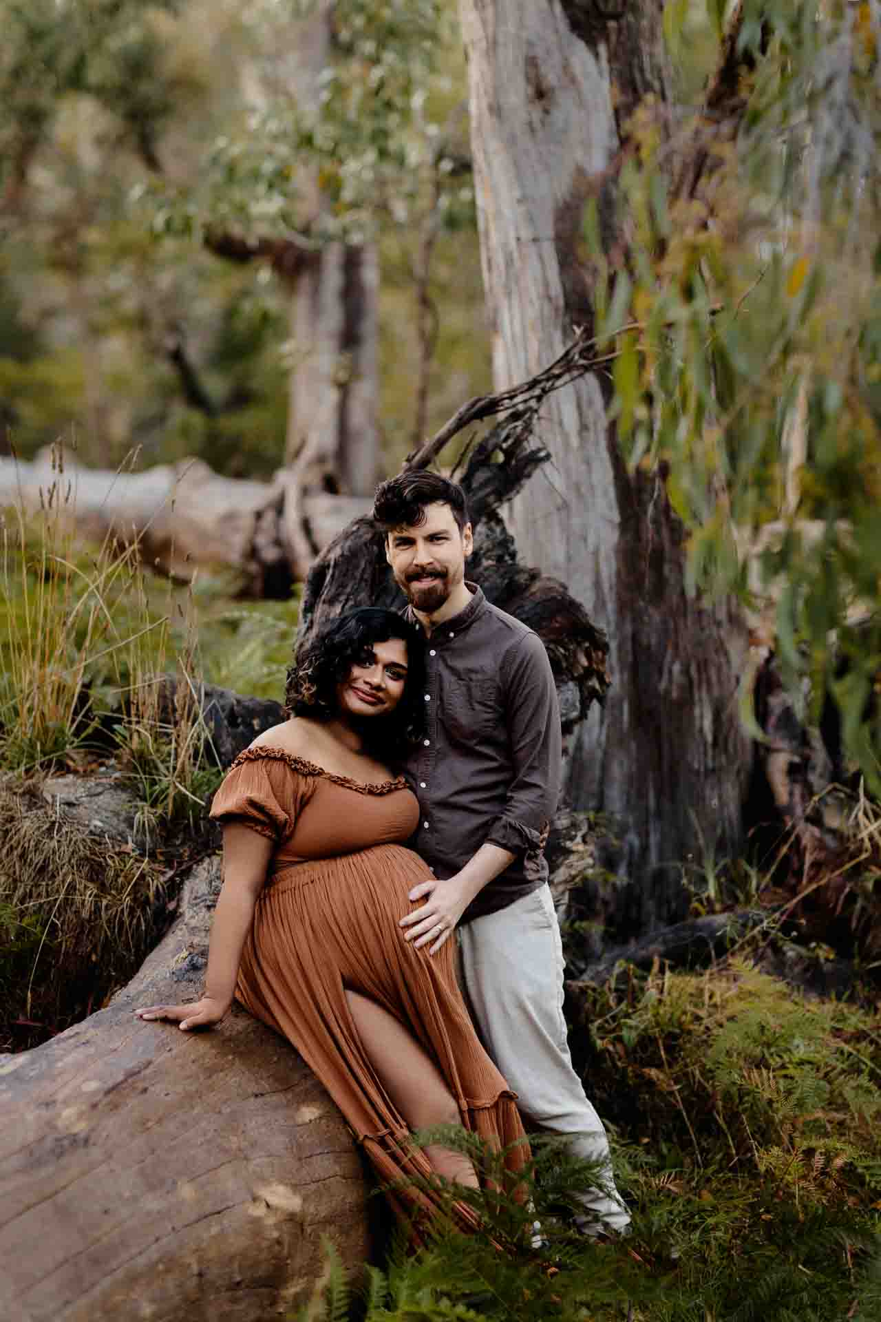 A pregnant mother wearing a rust dress sitting on a fallen tree with her partner at a maternity session in the Adelaide Hills.