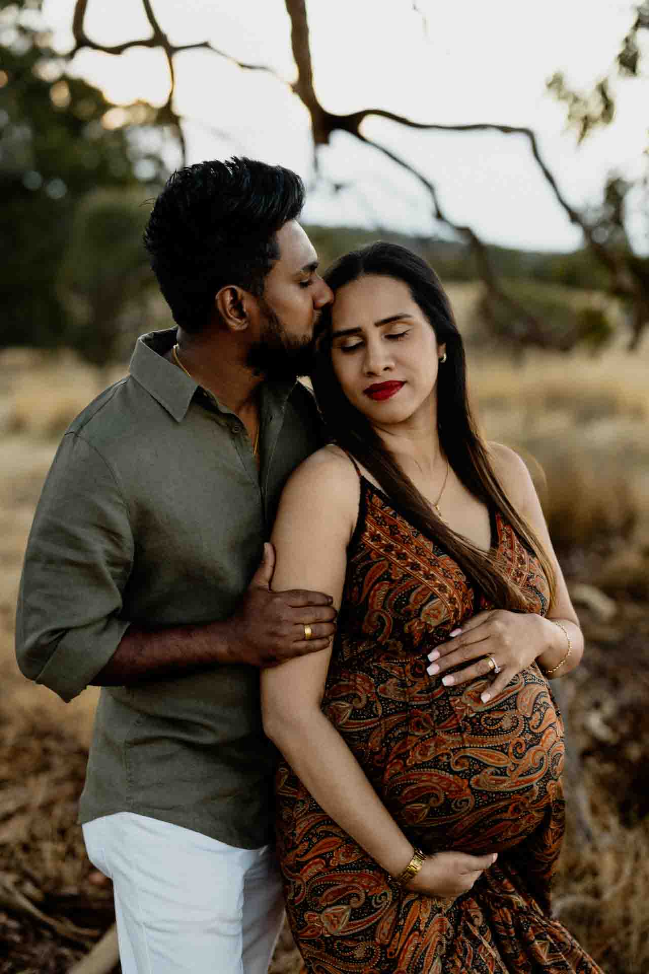 A man lovingly kisses a pregnant woman on the forehead as they embrace outdoors. The woman cradles her baby bump with both hands, and both appear calm and content in a natural, rustic setting—perfect for Adelaide baby photos.