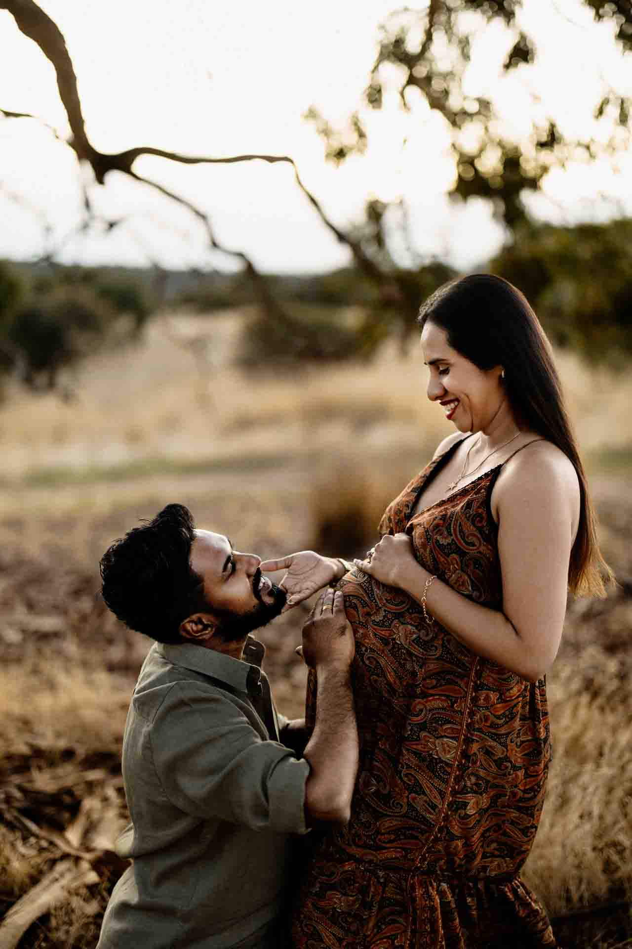 A man kneels and lovingly touches the pregnant belly of a smiling woman standing outdoors in a field, both sharing a joyful, intimate moment—perfect for Adelaide baby photos.