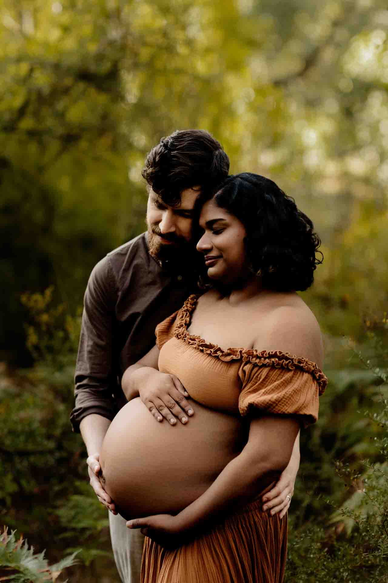 A pregnant mother wearing a rust dress cuddling with her partner at a maternity session in the Adelaide Hills.