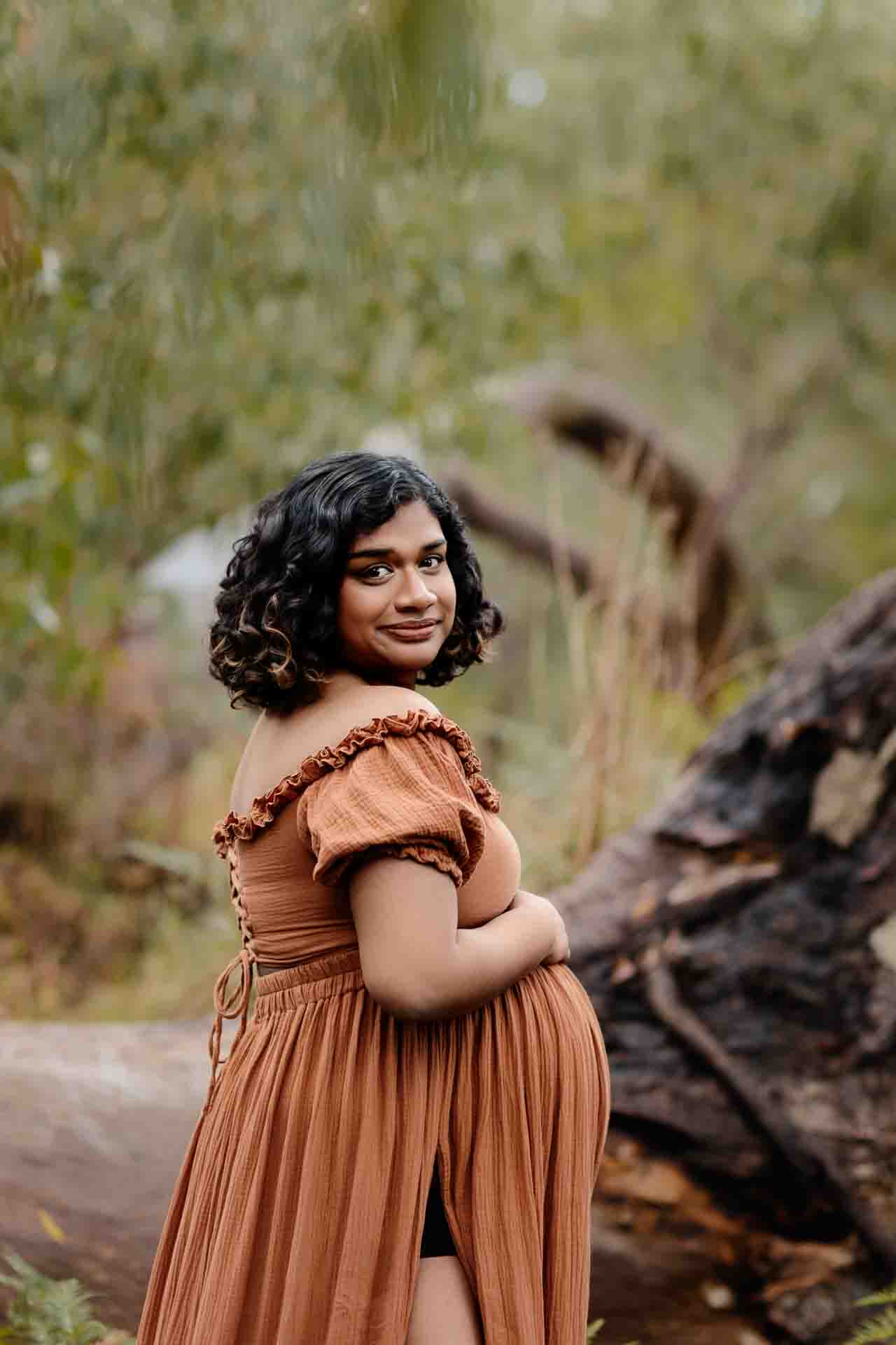 A pregnant mother wearing a rust dress looking over her shoulder at a maternity session in the Adelaide Hills.