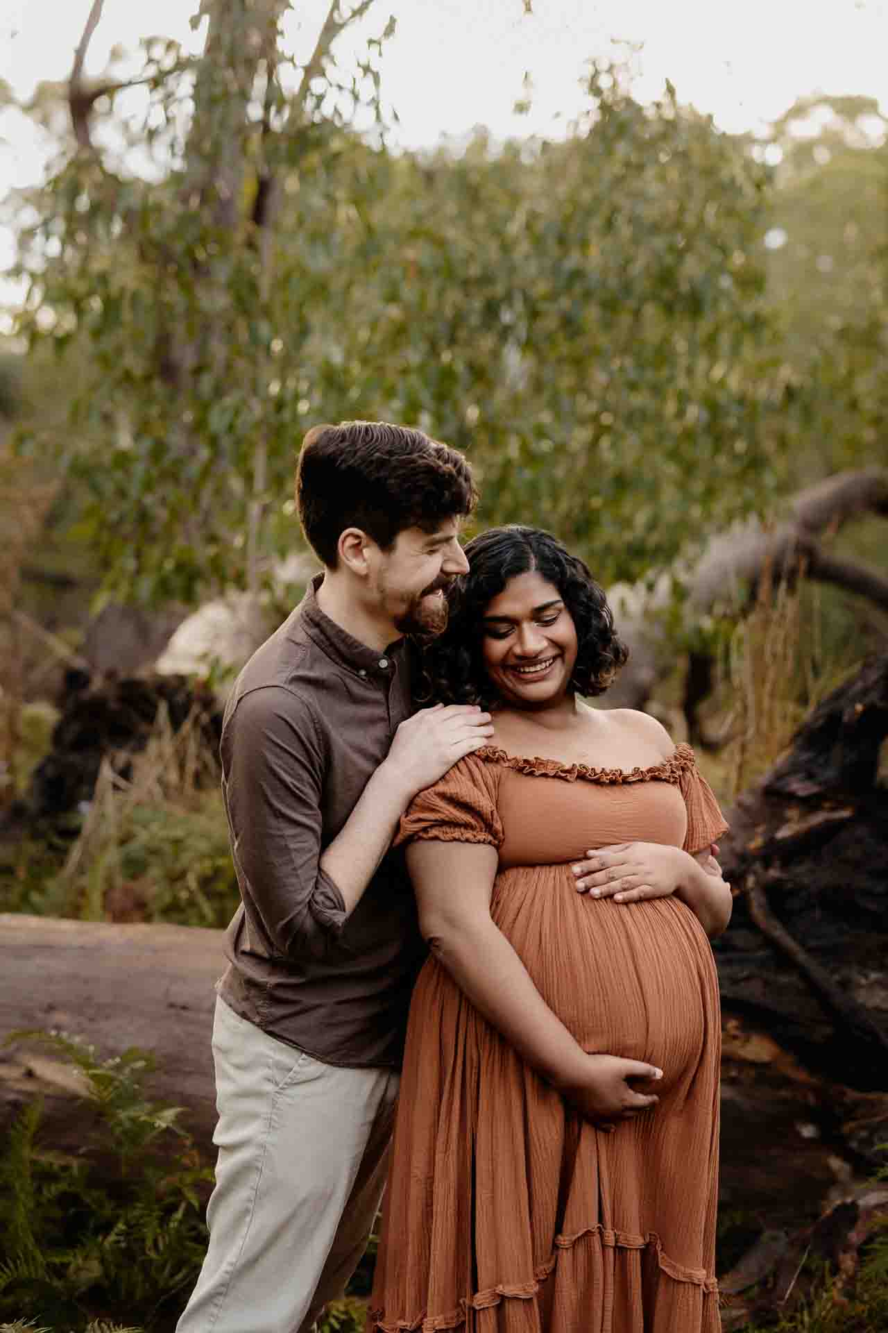 A pregnant mother wearing a rust dress laughing with her partner at a maternity session in the Adelaide Hills.