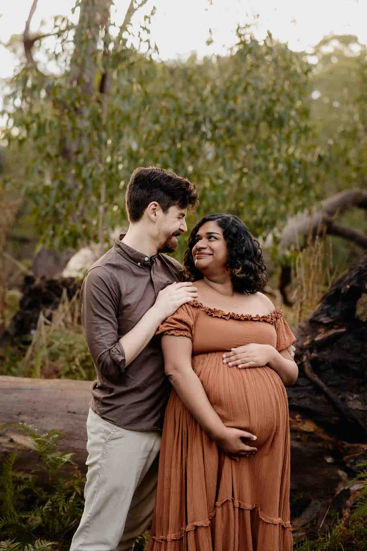 A pregnant mother wearing a rust dress cuddling looking at her partner at a maternity session in the Adelaide Hills.