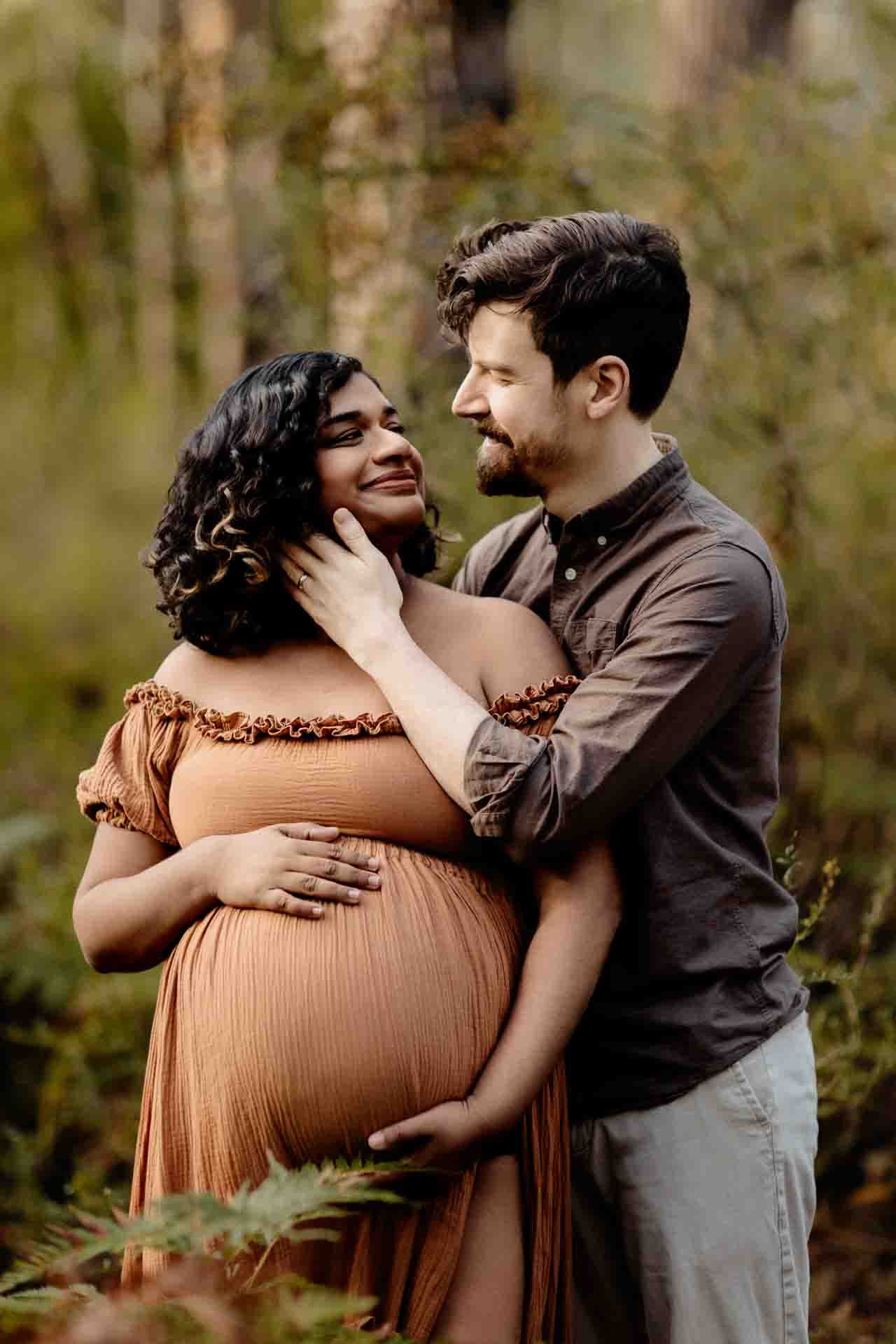 A pregnant mother wearing a rust dress snuggling with her partner at a maternity session in the Adelaide Hills.