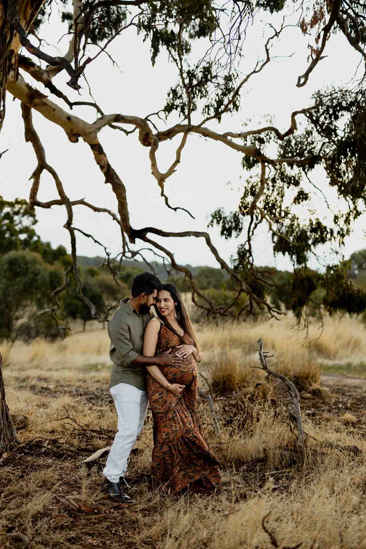A couple stand close together under a large, twisting tree in a grassy, dry landscape. The man embraces the smiling woman from behind as they share an affectionate moment, reminiscent of joyful Adelaide baby photos.