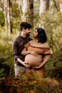 A pregnant mother wearing a rust dress with her pregnant belly out cuddling with her partner at a maternity session in the Adelaide Hills.