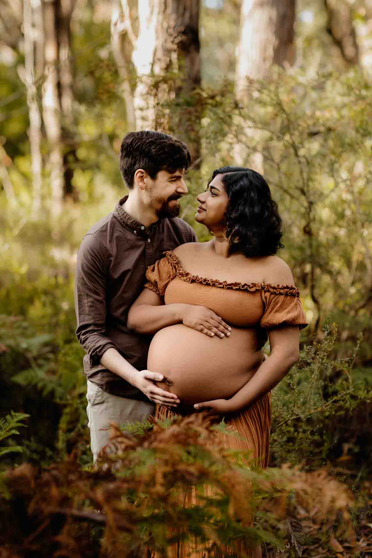 A pregnant mother wearing a rust dress with her pregnant belly out cuddling with her partner at a maternity session in the Adelaide Hills.