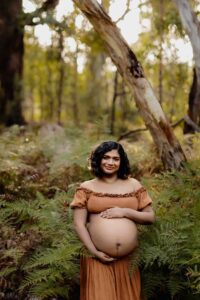 A pregnant mother wearing a rust dress smiling while being surrounded by ferns at a maternity session in the Adelaide Hills.