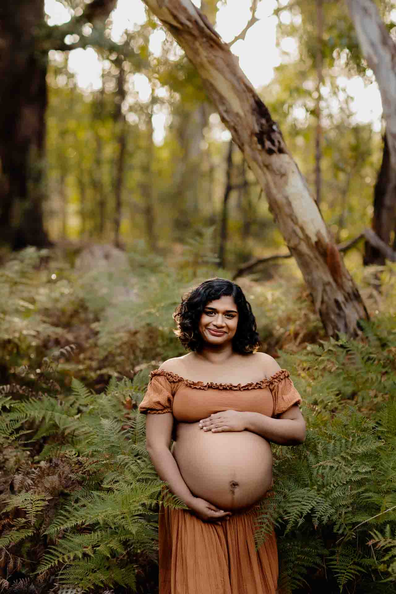 A pregnant mother wearing a rust dress smiling while being surrounded by ferns at a maternity session in the Adelaide Hills.