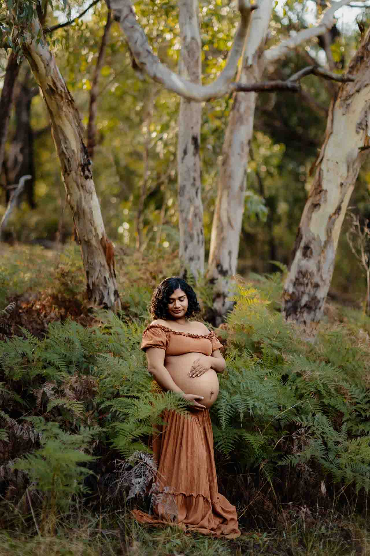 A pregnant mother wearing a rust dress surrounded by ferns at a maternity session in the Adelaide Hills.