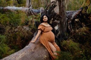 A pregnant mother wearing a rust dress sitting on a fallen tree at a maternity session in Adelaide.