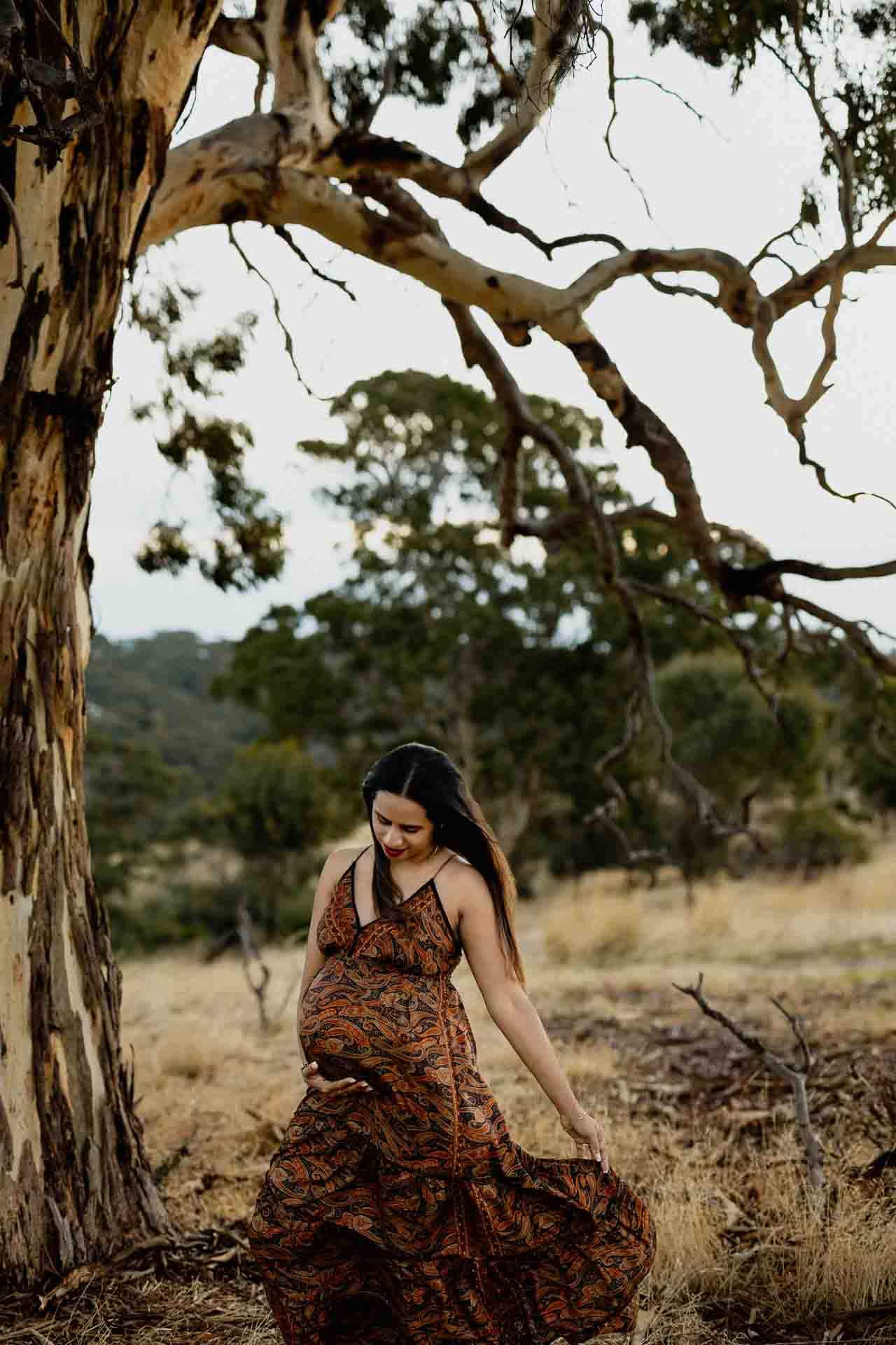 A pregnant woman in a patterned dress stands outdoors near a large tree, gently holding her dress. The natural, wooded setting is perfect for Adelaide baby photos, with dry grass and trees creating a serene backdrop.