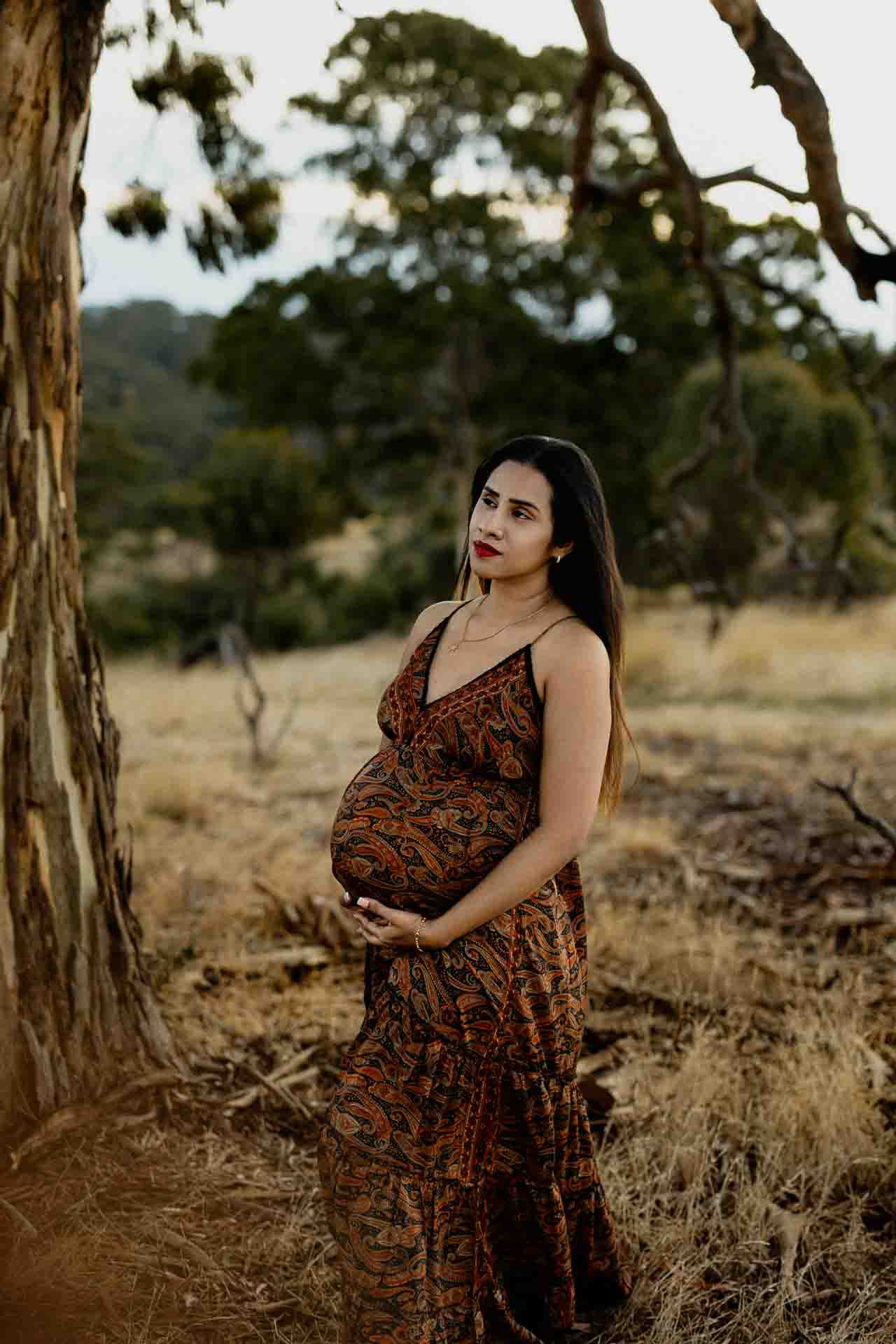 A pregnant woman in a patterned dress stands outdoors by a tree, gently holding her bump and looking thoughtfully into the distance—a serene moment perfect for Adelaide baby photos, with dry grass and trees softly framing the scene.