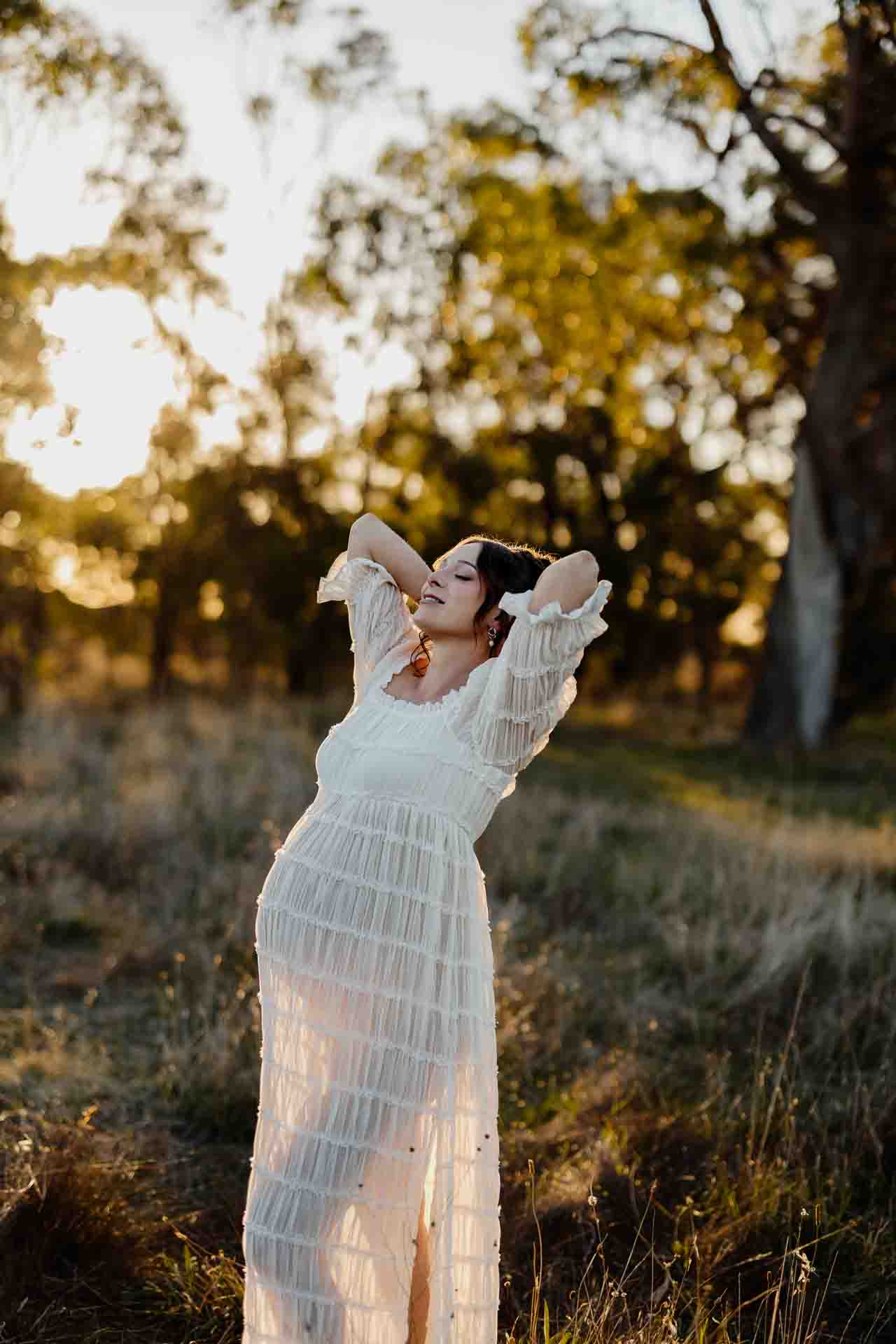 A pregnant mother in a white dress with her hands behind her head at a maternity session in Adelaide.