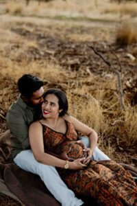 A couple sits on a blanket in a dry grassy field. The man embraces the woman from behind as she smiles, resting her hands on her pregnant belly, forming a heart—an adorable moment captured in these Adelaide baby photographs.