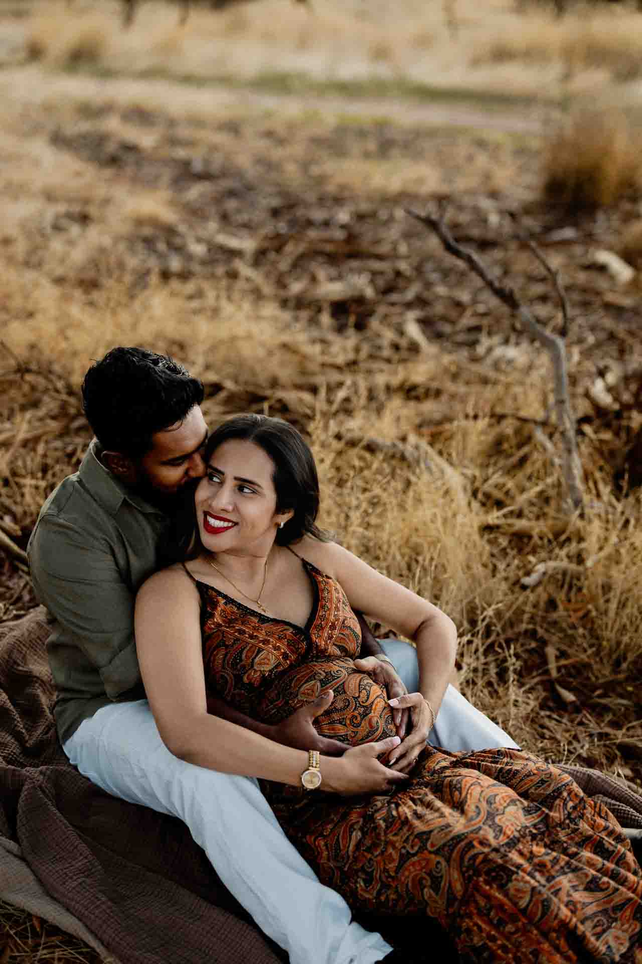 A couple sits on a blanket in a dry grassy field. The man embraces the woman from behind as she smiles, resting her hands on her pregnant belly, forming a heart—an adorable moment captured in these Adelaide baby photographs.