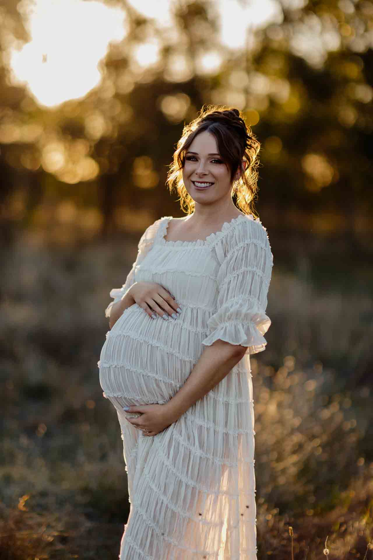 A pregnant mother in a white dress smiling and holding her pregnant belly at a maternity session in Adelaide.