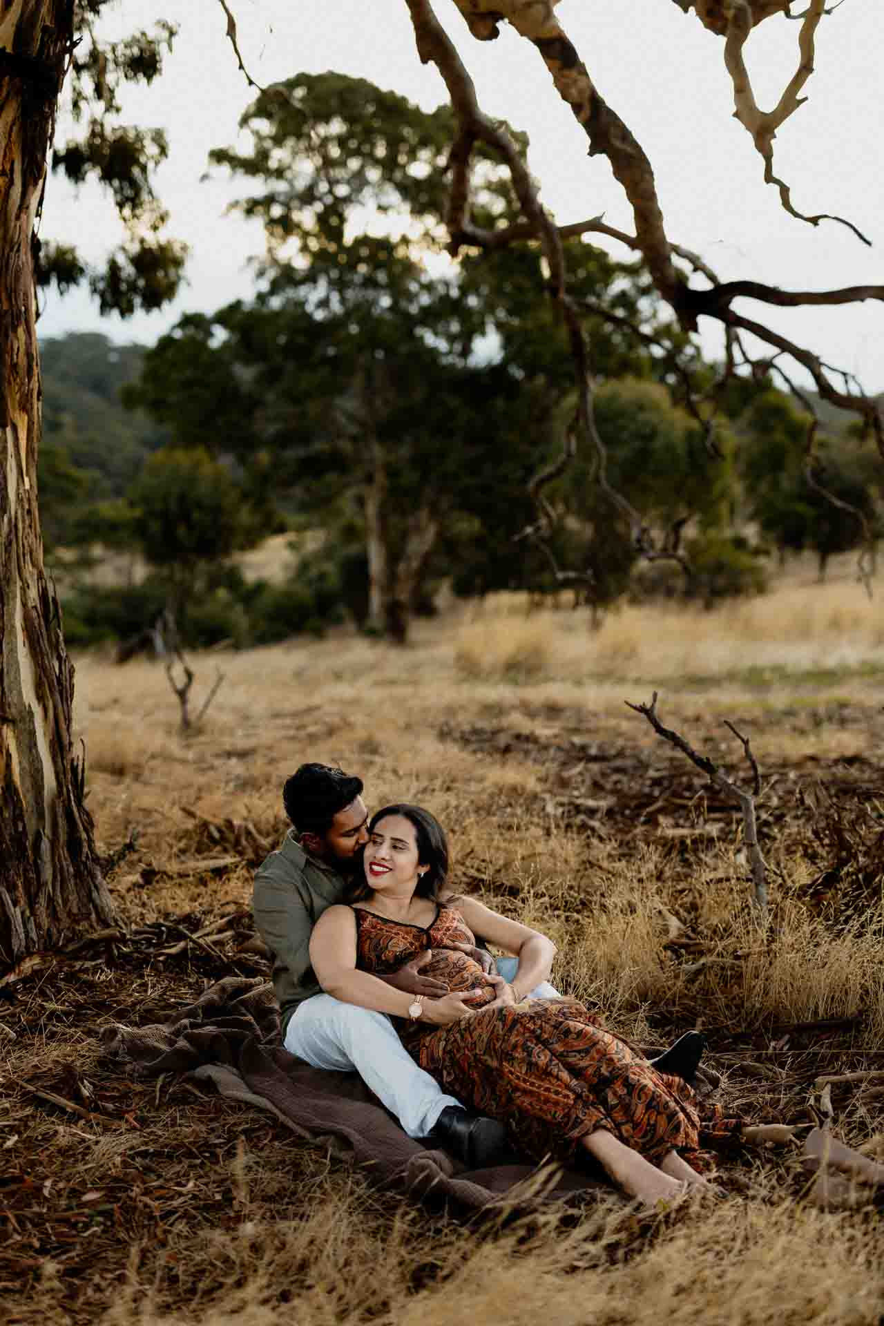 A couple sits on a blanket under a large tree in a grassy, wooded area. The woman, wearing a patterned dress, leans back and smiles at the man who sits behind her, embracing her affectionately—capturing a tender moment reminiscent of Adelaide baby photos.