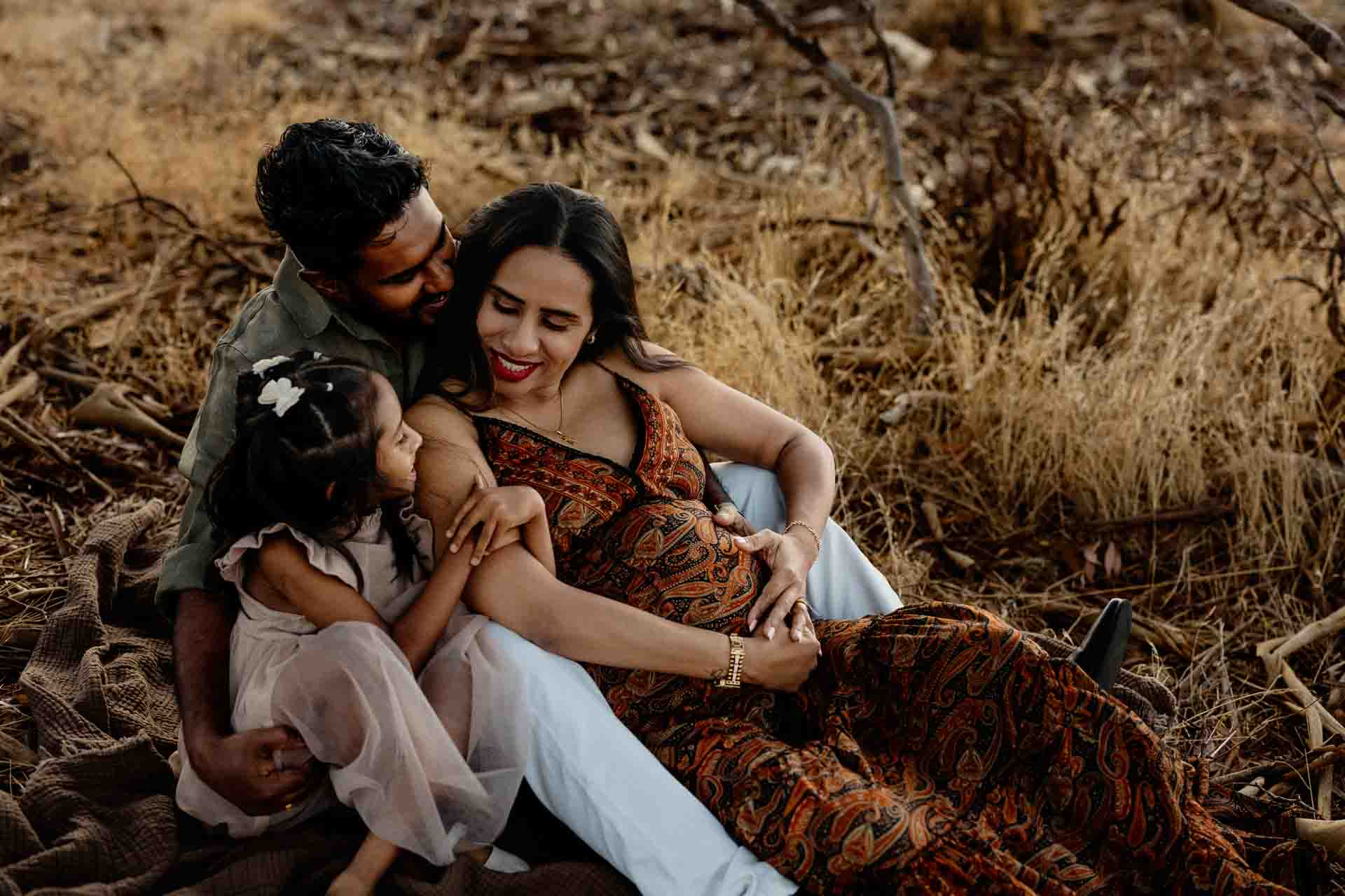 A family sits closely together outdoors on dry grass. The father hugs the mother, who is pregnant and wearing a patterned dress, whilst their young daughter sits on her lap, looking at her parents lovingly—capturing precious Adelaide baby photos.