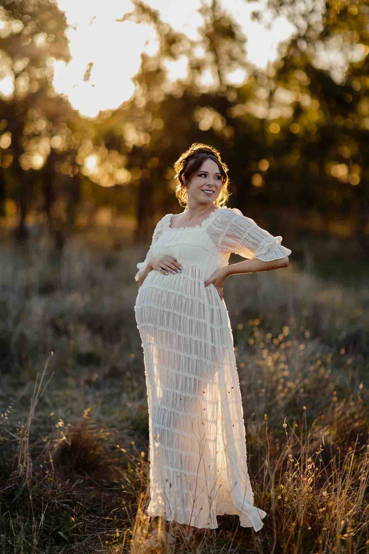 A pregnant mother in a white dress smiling and looking to the right at a maternity session in Adelaide.