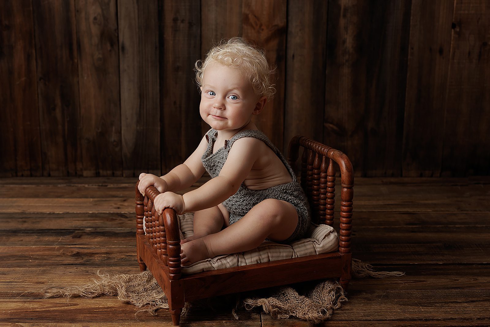 A smiling baby with curly blonde hair sits on a small wooden bed in this Adelaide baby photoshoot. Wearing a grey outfit, the little one is surrounded by rustic wooden planks, creating a warm, cosy setting.