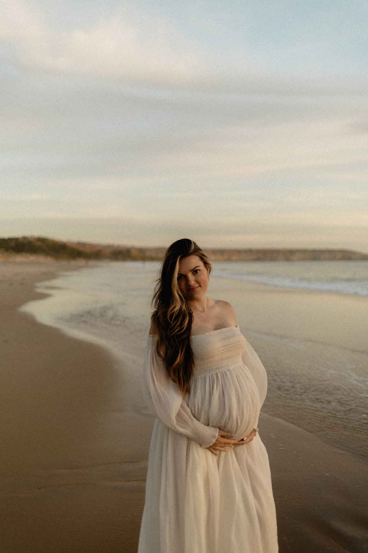 Pregnant woman standing on the shoreline in a flowing white dress