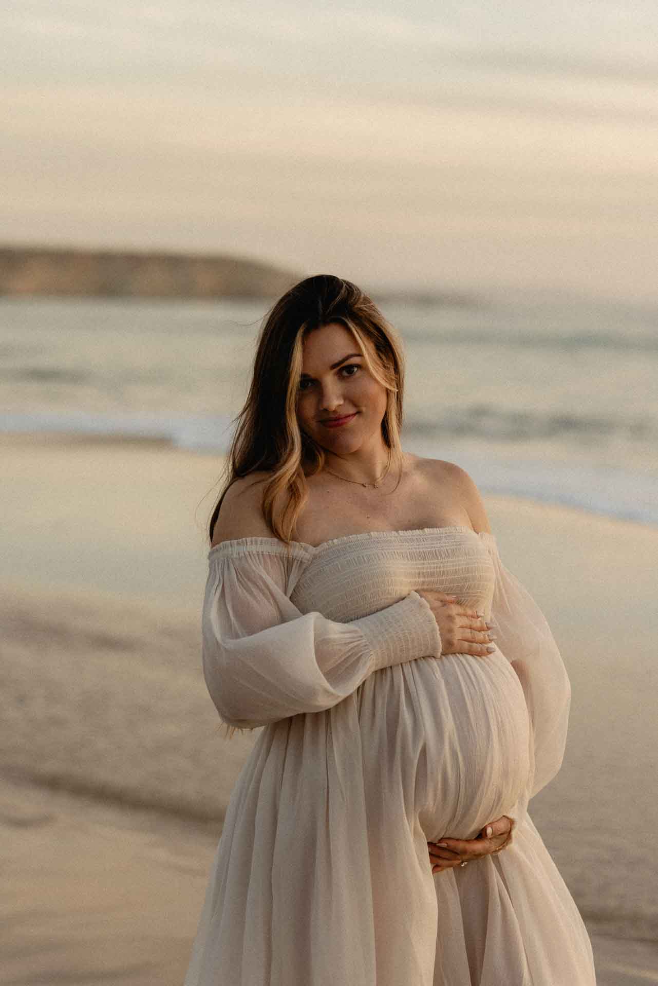 Pregnant woman posing on the beach in a flowing white maternity dress
