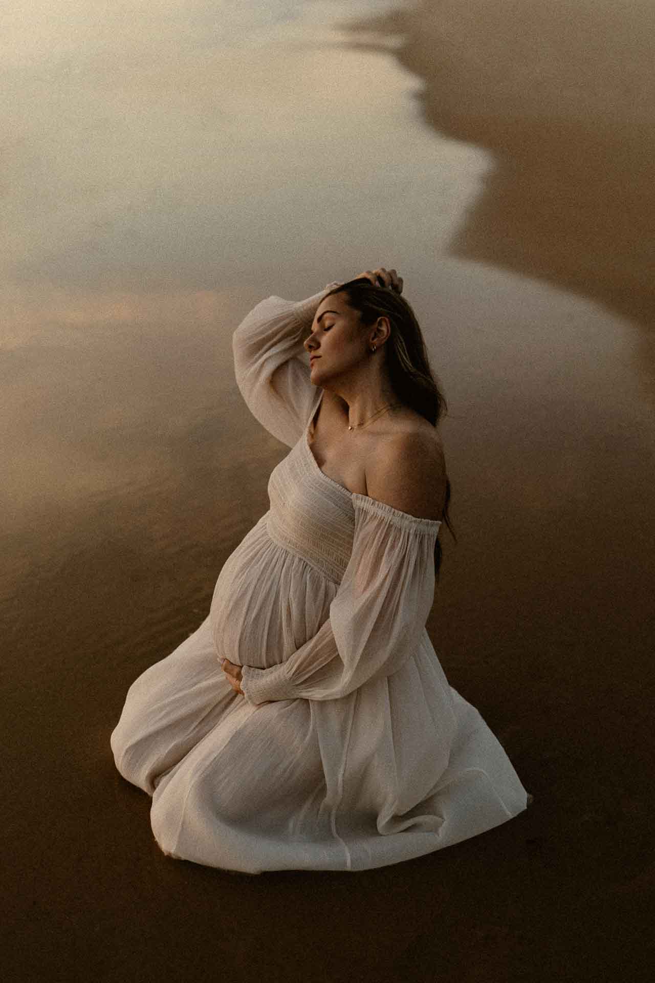 Pregnant woman kneeling on the wet sand at sunset in a flowing white dress