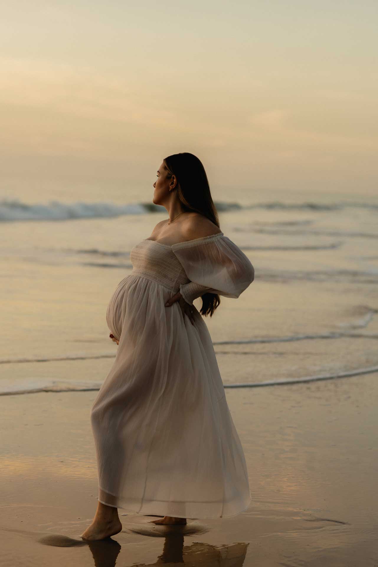 Pregnant woman standing in the ocean at sunset wearing a flowing white dress