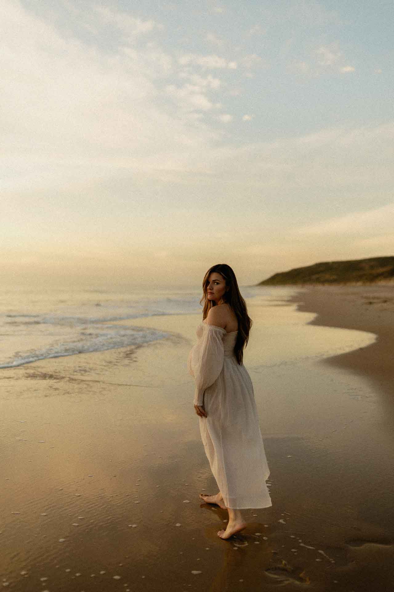 Pregnant woman walking barefoot along the shoreline in a flowing white dress