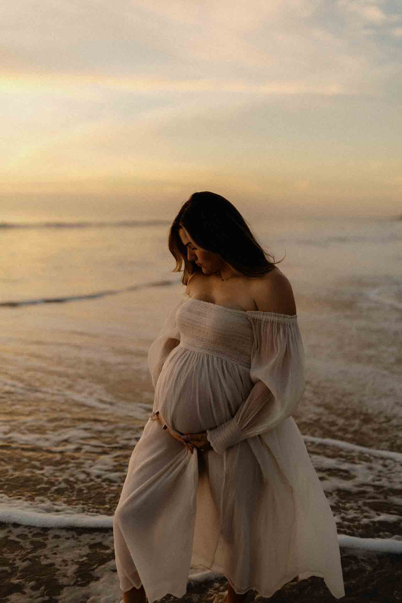 Pregnant woman standing beside the ocean in a flowing white maternity dress