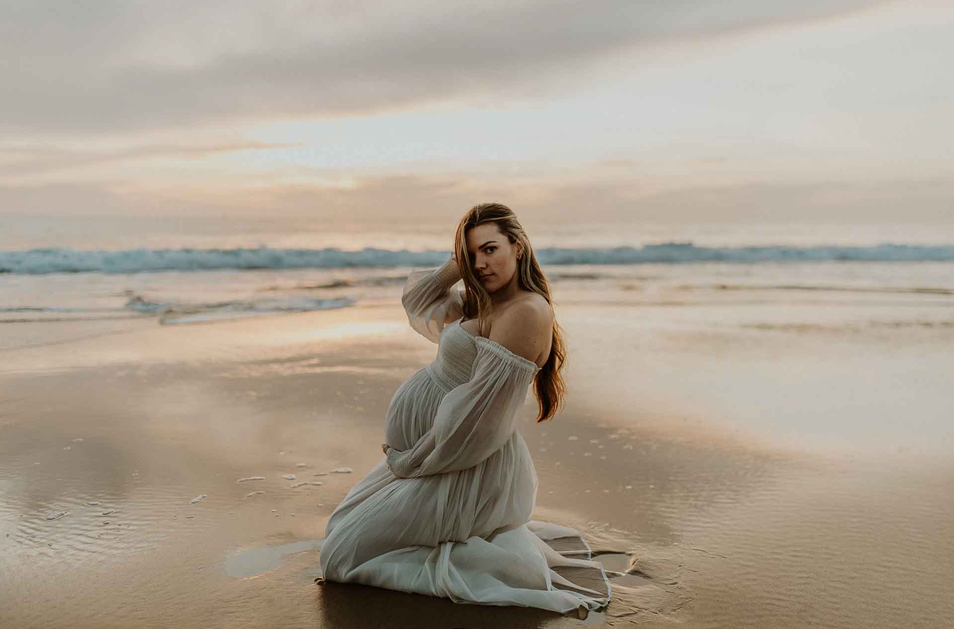 Pregnant woman kneeling beside the ocean in a flowing white dress